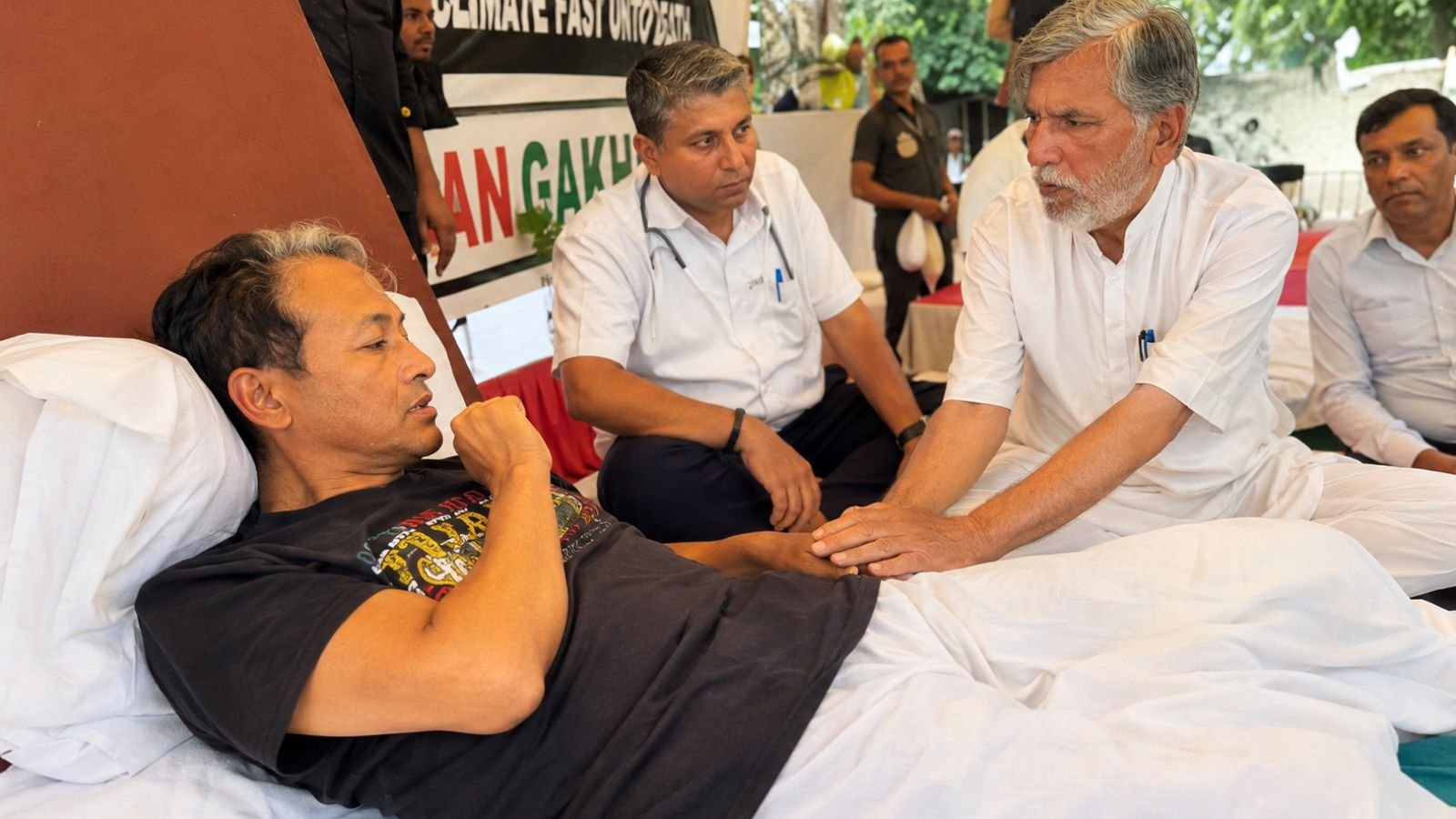 Sonam Wangchuk resting during a hunger strike at a protest site in New Delhi, surrounded by supporters and a medical professional as they discuss his health condition inside a peaceful protest camp.